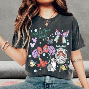 May include: A charcoal gray short-sleeve t-shirt featuring a graphic print with the text Part Of Your World. The design includes a mermaid, a crab, a fish, a dog, a bow, and seashell-shaped ears, all rendered in pastel colors.