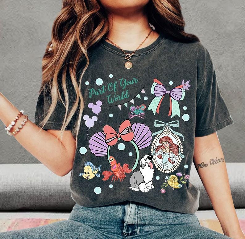 May include: A charcoal gray short-sleeve t-shirt featuring a graphic print with the text Part Of Your World. The design includes a mermaid, a crab, a fish, a dog, a bow, and seashell-shaped ears, all rendered in pastel colors.