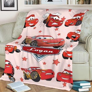 May include: A light pink fleece blanket featuring multiple images of the animated character Lightning McQueen from the movie Cars. The blanket has red star accents and the name "Logan" on a red and silver emblem.