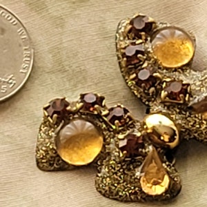 May include: A pair of gold tone butterfly shaped brooches with brown and clear rhinestones. The brooches have a textured surface and are accented with gold tone beads.