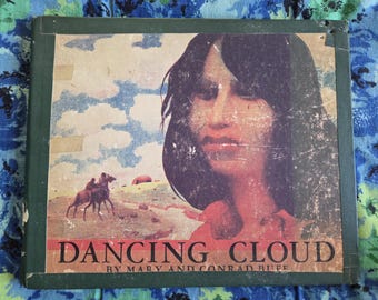 May include: A vintage book cover with a green border and a colorful illustration of a woman with long black hair. The title of the book is "Dancing Cloud" by Mary and Conrad Buff.