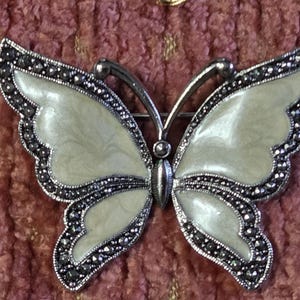 May include: A silver-toned butterfly brooch with cream-colored wings and a border of small, dark gray stones. The brooch has a pin on the back for secure attachment. The butterfly's wings are detailed with a subtle pattern.
