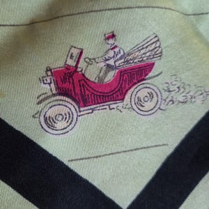 May include: Light green fabric with a repeating pattern of a vintage red car with a driver. The car has white wheels and is emitting smoke. Black diagonal stripes frame the design.