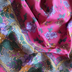 May include: A vibrant pink scarf with a floral pattern. The scarf features a repeating design of purple, yellow, and green flowers on a pink background. The border has a geometric pattern with a pink trim.