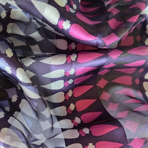 May include: A close-up of a flowing fabric with a repeating pattern of white, pink, and purple teardrop shapes. The fabric has a silky sheen and is draped in soft folds, creating a dynamic visual texture. The background is a deep purple.