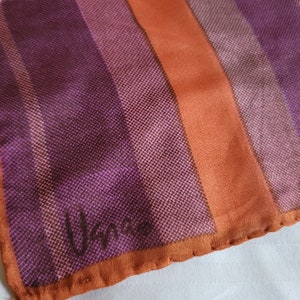 May include: A silk scarf with a striped pattern in shades of purple, orange, and tan. The scarf has a textured appearance and a brown border. The word "Vance" is printed in the corner.
