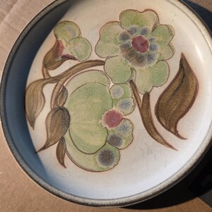 May include: A round ceramic plate with a floral design. The plate features a cream-colored background with a brown rim. The floral design includes green, brown, and red accents. The plate is likely used for serving food.
