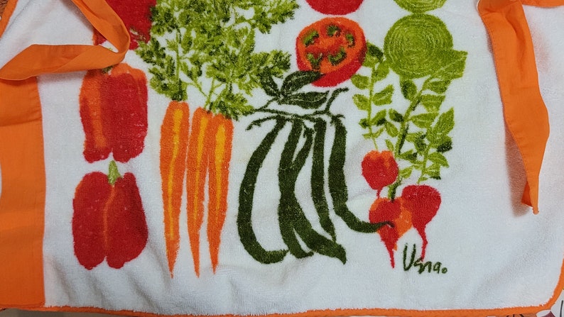 K&ouml;nnte beinhalten: Wei&szlig;es K&uuml;chentuch mit orangefarbenem Rand und einem bunten Gem&uuml;seprint. Der Druck zeigt Karotten, Paprika, Tomaten, gr&uuml;ne Bohnen und Radieschen. Das Handtuch hat das Wort "Usna" in der unteren rechten Ecke gedruckt.