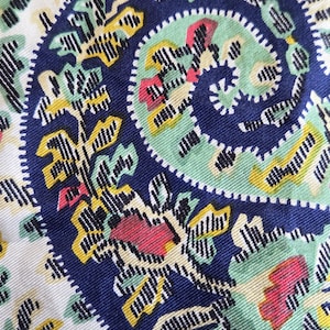May include: Close-up of a fabric with a paisley design. The pattern features a navy blue spiral in the center, surrounded by white, green, yellow, and red floral and leaf motifs. The fabric has a textured appearance.