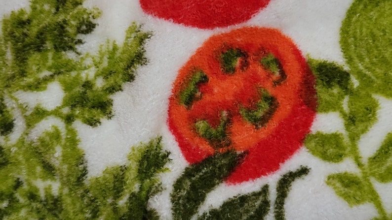 K&ouml;nnte beinhalten: Nahaufnahme eines wei&szlig;en Stoffes mit einer roten Tomate und gr&uuml;nen Blattmotiven. Die Tomate ist flauschig und hat eine rote und orange Farbe.