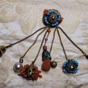 May include: An ornate vintage brooch with a central filigree design, adorned with turquoise beads and coral accents. Dangling from the brooch are chains with a turquoise bell, a faux pearl, and coral clusters. The brooch has a gold-tone finish.