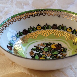 May include: A small white ceramic bowl with a green and gold floral design. The bowl has a gold rim and a green base.