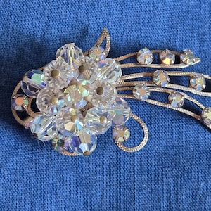 May include: A vintage brooch with a floral design. The brooch features clear, iridescent crystal beads clustered in a flower shape, with additional crystals set along silver-toned, curved accents. The brooch is set against a blue background.