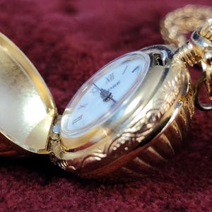 May include: A gold-tone pocket watch with a white face and black hands. The watch is attached to a gold-tone chain and has a decorative, ornate design.
