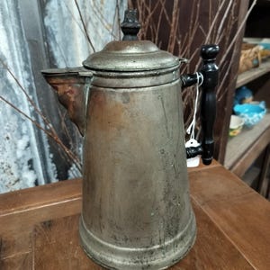 May include: A vintage silver and copper coffee pot with a black wooden handle. The pot has a worn, aged look and is sitting on a wooden surface.