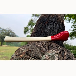May include: A wooden golf club with a cream-colored shaft and a bright red club head. The club is resting on a textured tree trunk, with a blurred green background of trees and sky.
