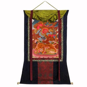 May include: A traditional Tibetan thangka painting on fabric. The artwork depicts a deity with a red face and blue body, riding a tiger. The background features clouds and other figures. The thangka is framed with a dark blue border and red ribbons.