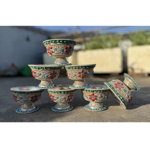 Puede incluir: Una colección de cuencos plateados ornamentados con intrincados diseños florales. Los cuencos están adornados con piedras rojas y turquesas y presentan una base de pedestal. Los cuencos están apilados y colocados sobre una superficie.
