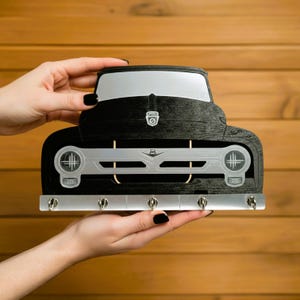 May include: A black wooden key holder in the shape of a classic truck. It features a silver-coloured grille and headlights, with hooks beneath. The key holder is held by hands with black nail polish.