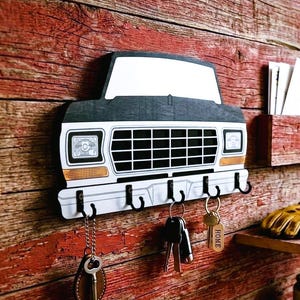 May include: A key holder in the shape of a vintage truck, mounted on a rustic red wooden wall. The key holder has a white grill, black hooks, and a small shelf. Keys hang from the hooks, adding a functional and decorative touch.