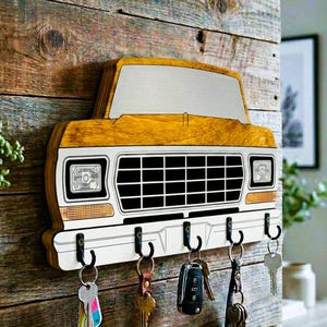 May include: A wooden key holder in the shape of a vintage truck front, painted yellow, white, and black. The key holder has six black hooks and is mounted on a rustic wooden wall. Several sets of keys hang from the hooks.