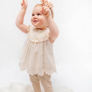 May include: A little girl wearing a cream-colored dress with lace trim and matching pants. She is wearing a headband with a flower on top. She is smiling and looking up.