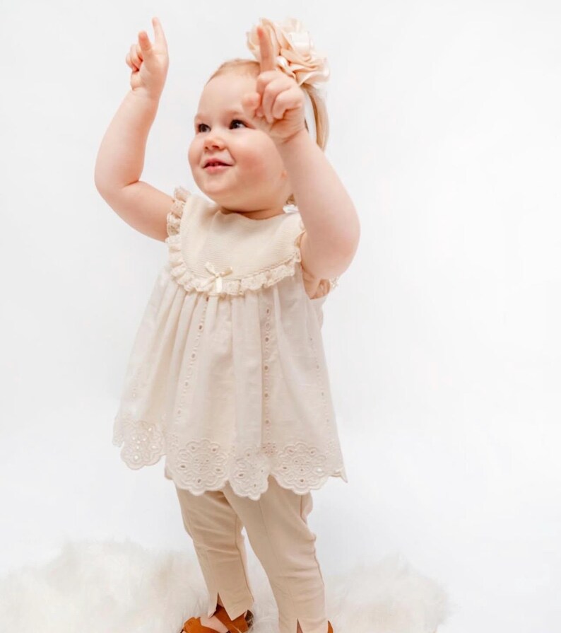 May include: A little girl wearing a cream-colored dress with lace trim and matching pants. She is wearing a headband with a flower on top. She is smiling and looking up.