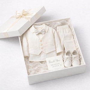May include: A cream-colored formal outfit set in a white gift box, including a long-sleeved shirt with a vest, a matching bow tie, trousers, and lace-up shoes. A small card inside reads: Thank You, Made with love for your little one.