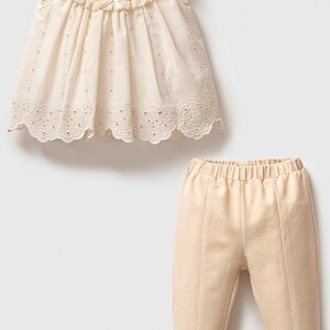 May include: A two-piece outfit for a baby or toddler, featuring a cream-colored sleeveless top with a lace trim and a matching pair of cream-colored pants.