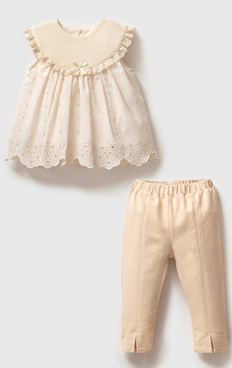 May include: A two-piece outfit for a baby or toddler, featuring a cream-colored sleeveless top with a lace trim and a matching pair of cream-colored pants.