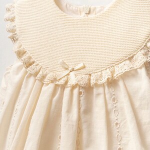 May include: A sleeveless, cream-colored dress with a lace trim and a crocheted collar. The dress has a bow at the neckline and a delicate lace pattern throughout.