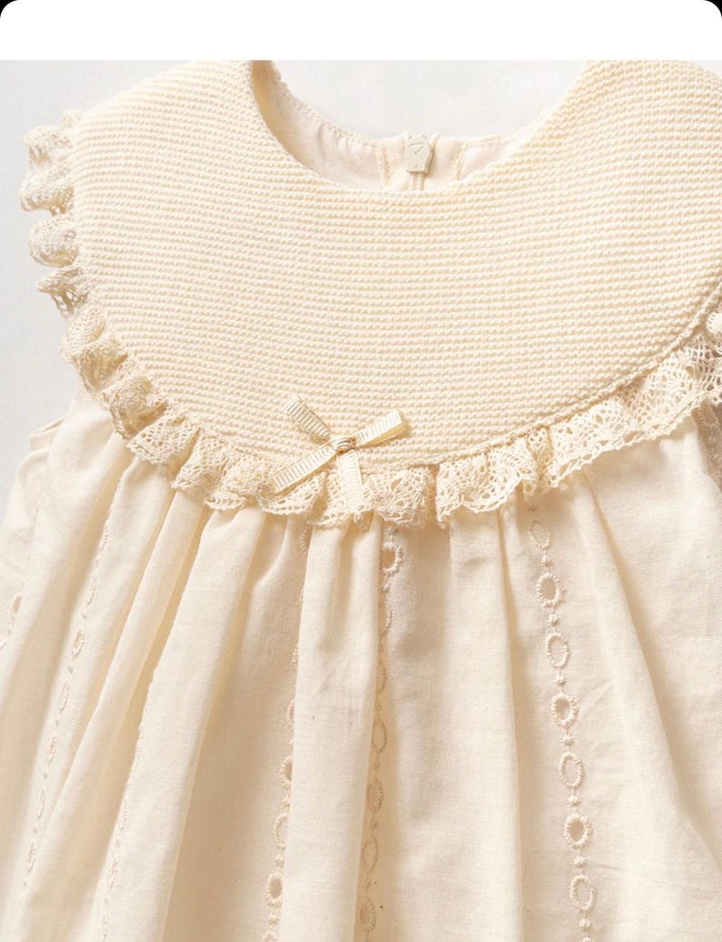May include: A sleeveless, cream-colored dress with a lace trim and a crocheted collar. The dress has a bow at the neckline and a delicate lace pattern throughout.