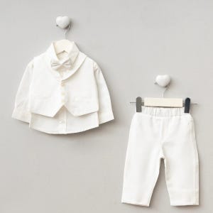 K&ouml;nnte beinhalten: Ein formelles wei&szlig;es Outfit f&uuml;r ein Baby oder Kleinkind, bestehend aus einem Hemd mit Kn&ouml;pfen, einer Weste, einer Fliege und einer Hose. Das Ensemble wird auf Kleiderb&uuml;geln vor einem hellgrauen Hintergrund pr&auml;sentiert, passend f&uuml;r einen besonderen Anlass.