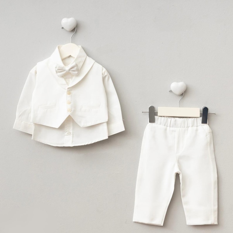 K&ouml;nnte beinhalten: Ein formelles wei&szlig;es Outfit f&uuml;r ein Baby oder Kleinkind, bestehend aus einem Hemd mit Kn&ouml;pfen, einer Weste, einer Fliege und einer Hose. Das Ensemble wird auf Kleiderb&uuml;geln vor einem hellgrauen Hintergrund pr&auml;sentiert, passend f&uuml;r einen besonderen Anlass.