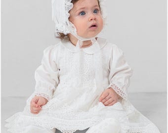 Puede incluir: Un bebé viste un vestido blanco de manga larga con detalles de encaje intrincados, cuello redondo y un gorro a juego con volantes delicados y tela de lunares. El conjunto incluye patucos blancos suaves que completan el atuendo coordinado.