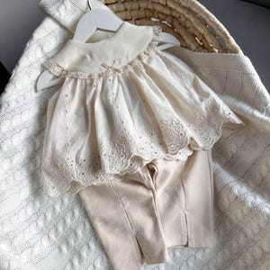 May include: A two-piece baby outfit in a cream color. The top has a lace trim and a peter pan collar. The pants are plain and have a straight leg.