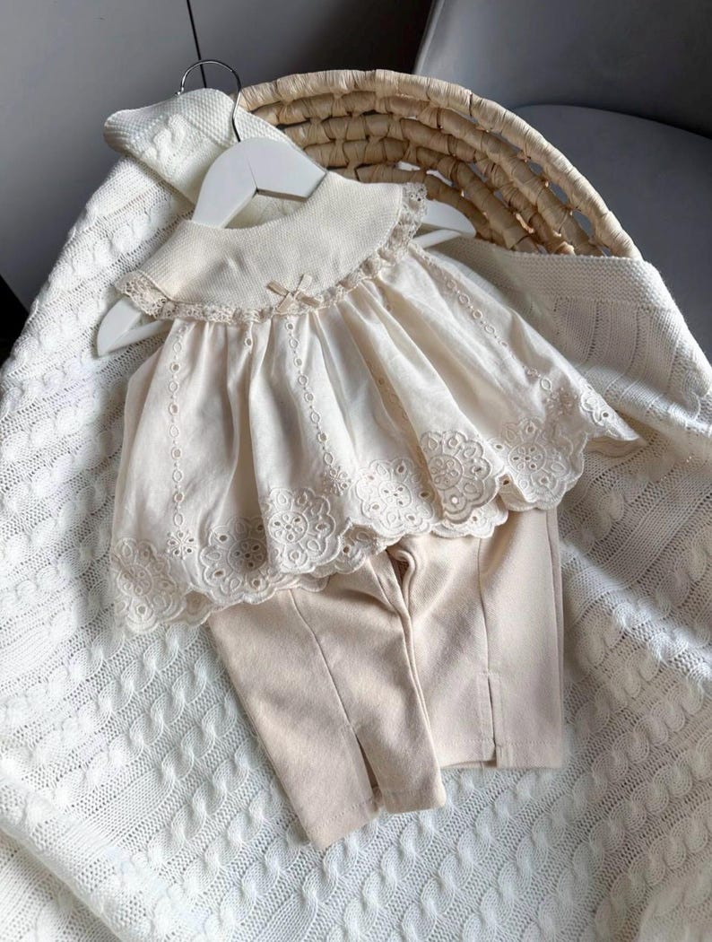 May include: A two-piece baby outfit in a cream color. The top has a lace trim and a peter pan collar. The pants are plain and have a straight leg.