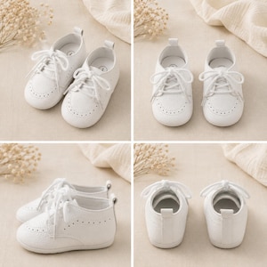 May include: A pair of white baby shoes with a textured pebble-grain finish, featuring a rounded toe, lace-up closure with white laces, and decorative perforated brogue detailing along the toe cap and sides, with a small pull tab at the heel.