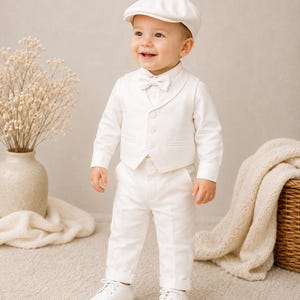 May include: A toddler wearing a white long-sleeved button-down shirt, a matching white vest with three buttons, white trousers with cuffed hems, a white bowtie, and a white flat cap. The outfit is completed with white leather dress shoes.