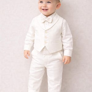 K&ouml;nnte beinhalten: Ein l&auml;chelndes Kleinkind in einem wei&szlig;en formellen Outfit, bestehend aus einem Hemd mit Kn&ouml;pfen, einer Weste, einer Hose, einer Fliege und einer M&uuml;tze. Das Outfit wird mit wei&szlig;en Turnschuhen kombiniert. Der Hintergrund ist eine helle, strukturierte Wand.
