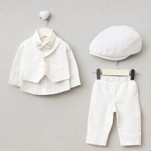 K&ouml;nnte beinhalten: Ein wei&szlig;es formelles Outfit f&uuml;r ein Baby oder Kleinkind, bestehend aus einem Hemd mit Weste, Fliege, Hose und einer Schieberm&uuml;tze. Die Kleidung wird auf Kleiderb&uuml;geln vor einem hellgrauen Hintergrund pr&auml;sentiert.