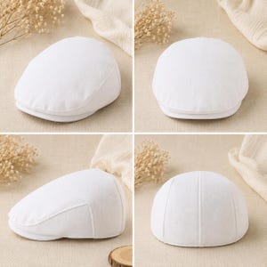 May include: A solid white flat cap featuring a structured crown and a short, stiff brim. The cap is constructed from a woven fabric with a visible texture, showcasing a classic rounded silhouette with stitched panel detailing on the back.
