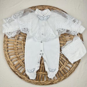 May include: A white long-sleeved footed romper featuring a sheer lace capelet overlay with scalloped edges. The garment includes a silver embroidered wing emblem at the collar and on each foot, with silver-toned snap buttons and a matching bonnet.