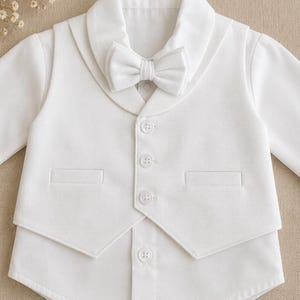 May include: A white long-sleeved shirt featuring an attached vest with a shawl collar and a matching textured bow tie. The vest is secured with three white buttons, and the shirt has a single visible button at the bottom hem with two pockets.