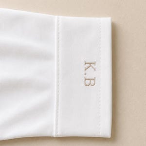 May include: A white long-sleeve shirt cuff featuring a structured, straight edge and a clean finish. The cuff is embroidered with the initials K.B in a light beige, serif-style font, positioned vertically on the right side of the fabric.