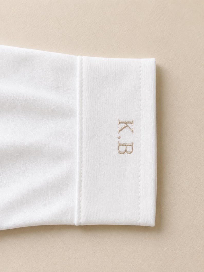 May include: A white long-sleeve shirt cuff featuring a structured, straight edge and a clean finish. The cuff is embroidered with the initials K.B in a light beige, serif-style font, positioned vertically on the right side of the fabric.