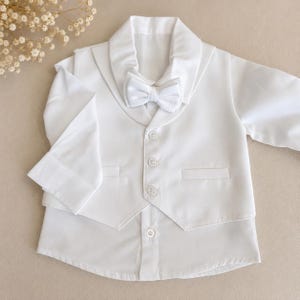 May include: A crisp white long-sleeved shirt featuring an attached vest with a shawl collar and three matching buttons. A textured white bow tie is secured at the neckline. The garment is constructed from a smooth, solid white fabric throughout.