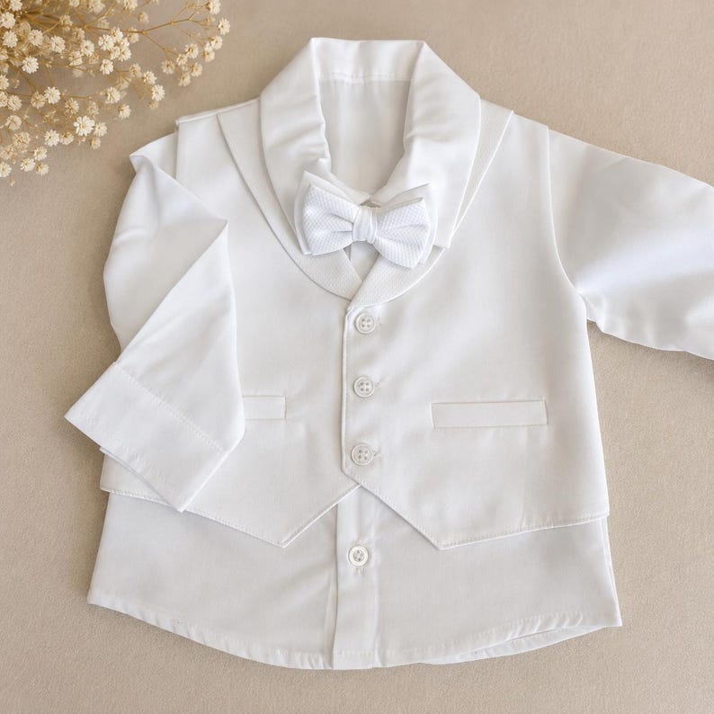 May include: A crisp white long-sleeved shirt featuring an attached vest with a shawl collar and three matching buttons. A textured white bow tie is secured at the neckline. The garment is constructed from a smooth, solid white fabric throughout.