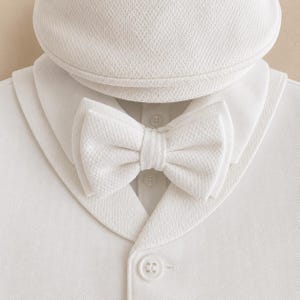 May include: A white textured fabric set consisting of a flat cap, a bow tie, and a collared shirt with a shawl lapel. The shirt features a central button placket with visible white buttons, and the bow tie is layered with a matching fabric texture.