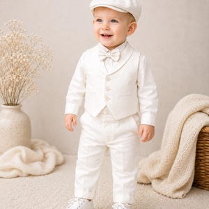 May include: A cream-colored formal outfit consisting of a long-sleeved button-down shirt, a matching vest with three buttons, trousers with cuffed hems, a bow tie, and a flat cap. The ensemble is completed with a pair of white lace-up shoes.